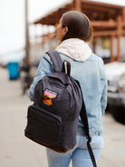 A woman wearing a denim jacket carries the Black Backpack w/ Outer Sunset Patch as she walks outdoors near buildings and parked cars.