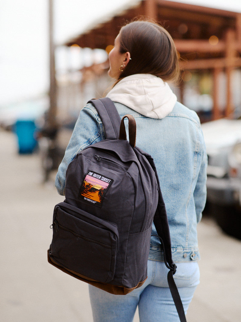 A woman wearing a denim jacket carries the Black Backpack w/ Outer Sunset Patch as she walks outdoors near buildings and parked cars.