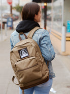 A person in a denim jacket strolls along the city sidewalk, showcasing a stylish Olive Backpack w/ SFP Patch with leather trims that captures attention effortlessly.
