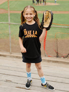 A smiling girl in the Youth Beat LA Tee - Black & Orange celebrates Bay Area sports, holding a baseball glove at a ballpark while standing on the bleachers.