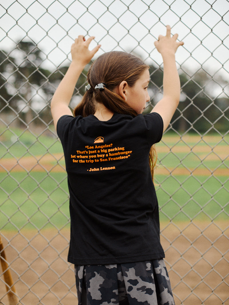 A girl with long brown hair stands at a chain-link fence, wearing the Youth Beat LA Tee - Black & Orange featuring bold yellow text on the back—a tribute to Bay Area sports pride.