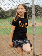 A young girl wearing the Youth Beat LA Tee - Black & Orange stands by a baseball field fence, smiling and holding a glove.
