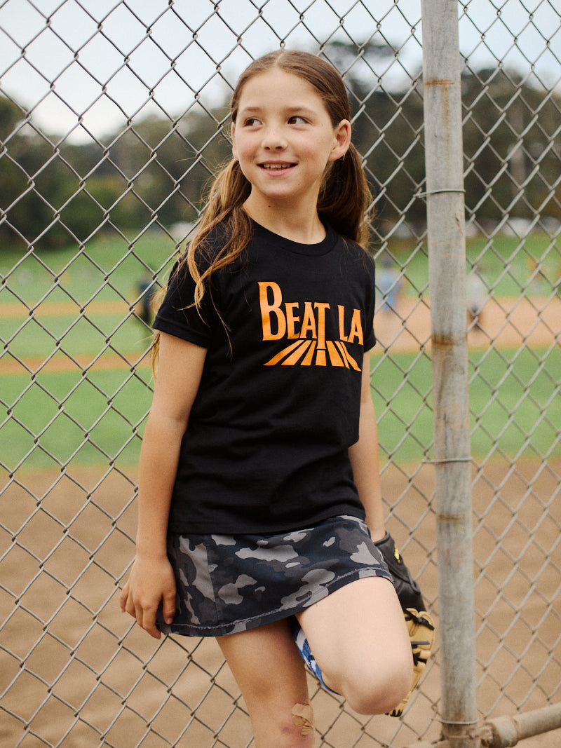 A young girl wearing the Youth Beat LA Tee - Black & Orange stands by a baseball field fence, smiling and holding a glove.