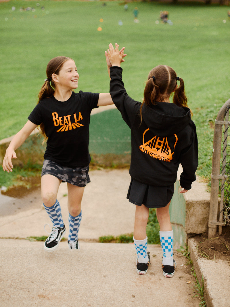 Two girls in sportswear, one wearing the Youth Beat LA Tee - Black & Orange, high-five each other outside on a concrete path near a grassy field.