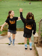 Two girls in matching socks and sportswear, including Youth Black & Orange Logo Zip-Up Hoodies, high-five on a park path.