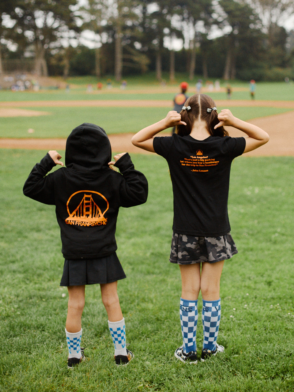 Two kids, each wearing a youth black & orange logo zip-up hoodie and blue checkered socks, stand on a grassy field near a baseball diamond.