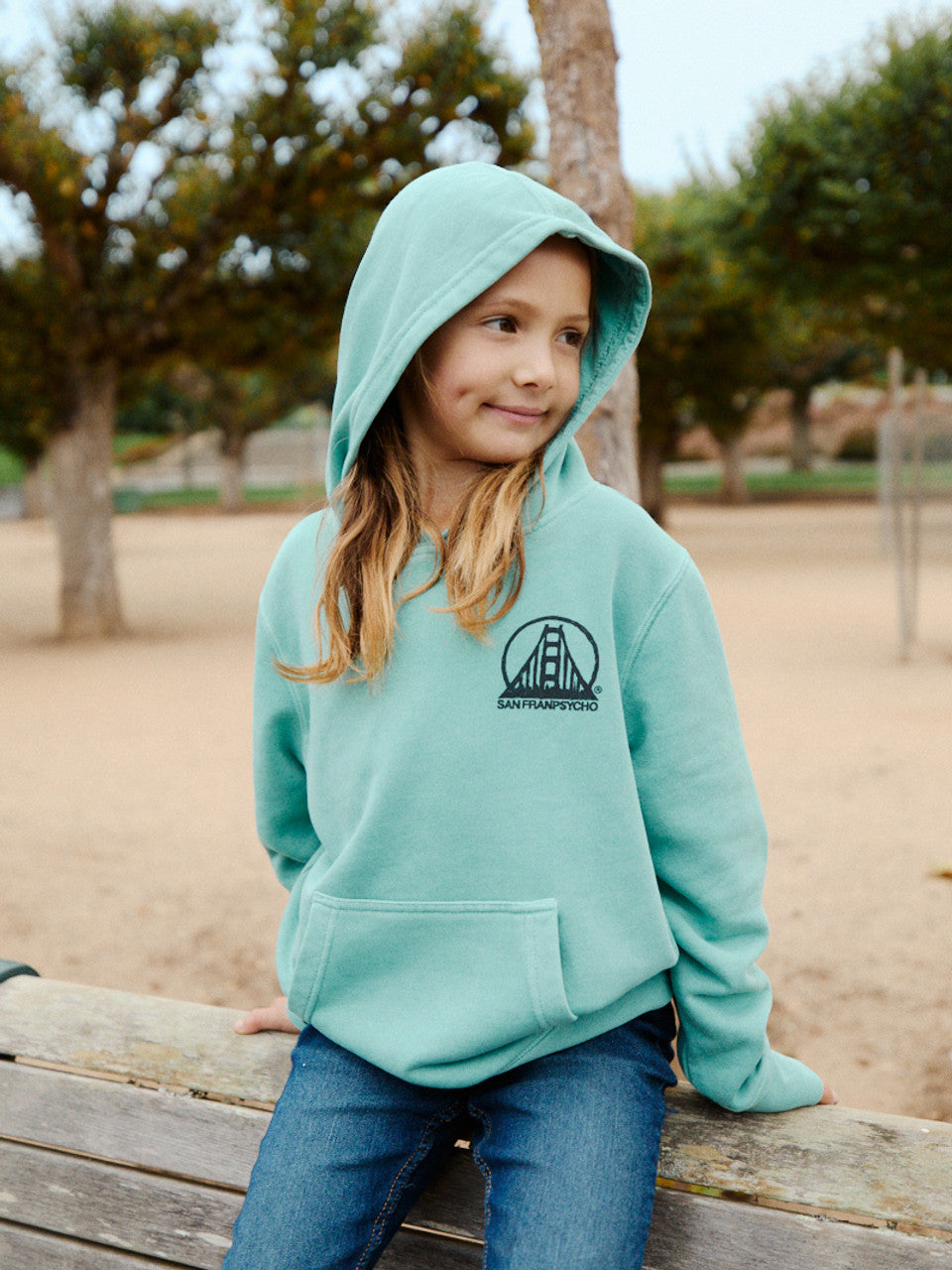 A young girl in a Youth Mint Scribble Logo Pullover Hoodie sits on a park bench. She's smiling warmly, with the lush trees framing her joyful moment.