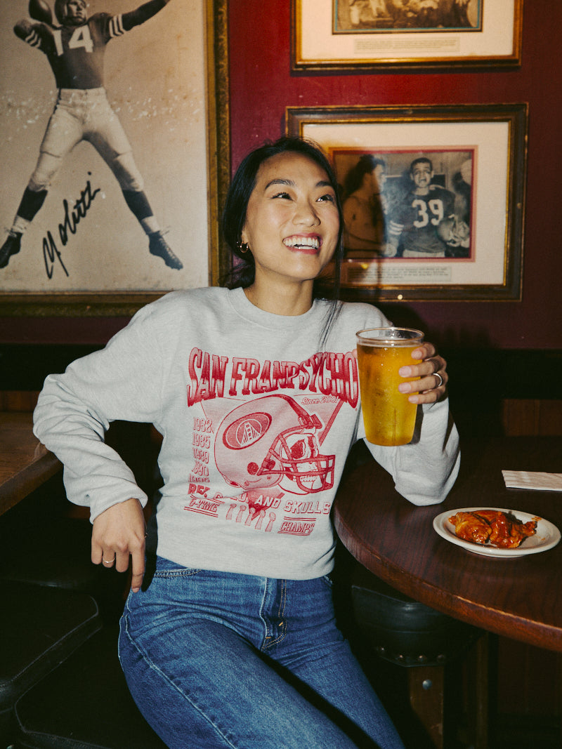 A person, dressed in a Candlestick Champs Crewneck, is laughing and holding a pint of beer in a lively sports bar adorned with framed photos on the wall.