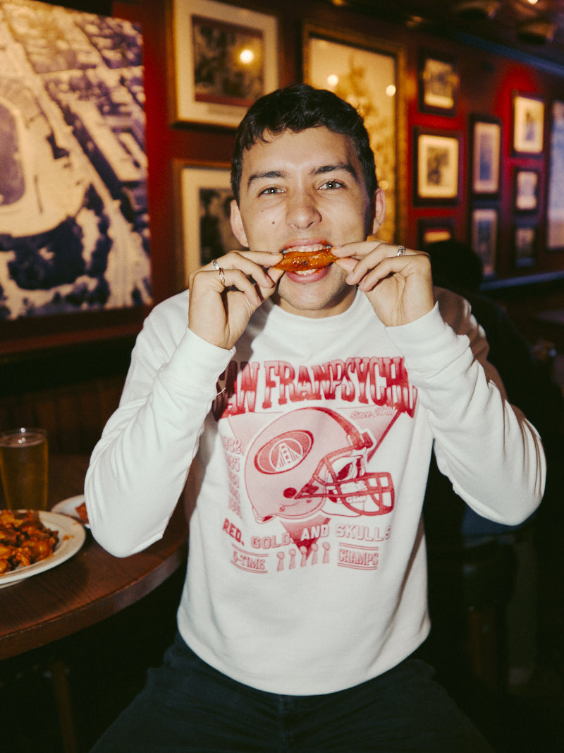 A person enjoys chicken wings in a restaurant while wearing the Candlestick Champs Crewneck, a white graphic vintage sweatshirt that captures the spirit of the Niners season.