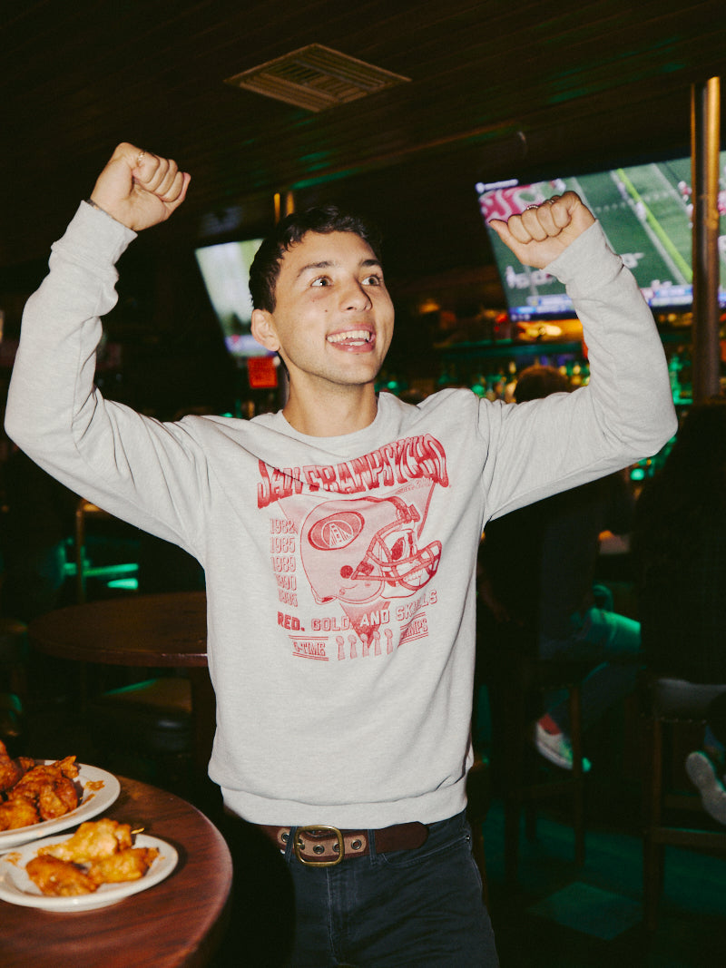 Wearing a Candlestick Champs Crewneck, someone cheers in a sports bar filled with TV screens and plates of food, celebrating the Niners season.