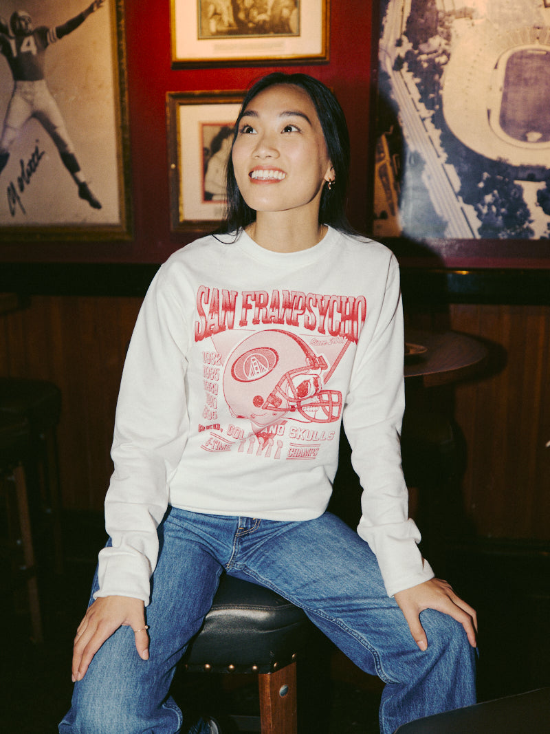 A person sits on a stool, smiling in a Candlestick Champs Crewneck adorned with San Francisco football graphics, capturing the energy of Niners season in a lively bar setting.
