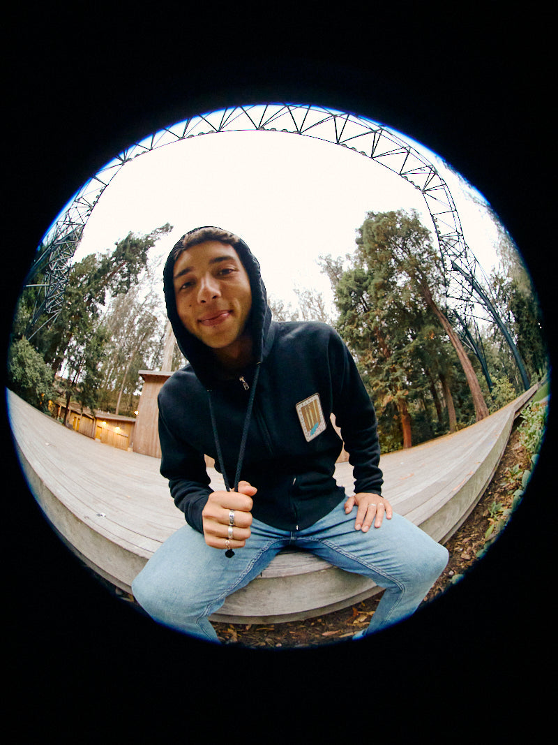 A person in a Black Muir Woods Patched Zip-Up Hoodie sits on a wooden platform surrounded by trees, all viewed through the intriguing distortion of a fisheye lens. Embracing the spirit of 'Free To Wander,' this scene captures a moment of quiet reflection in nature.