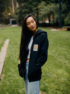 A person with long hair, wearing a Black Muir Woods Patched Zip-Up Hoodie and jeans, stands outdoors on a grass path. They seem free to wander and embrace the natural surroundings.