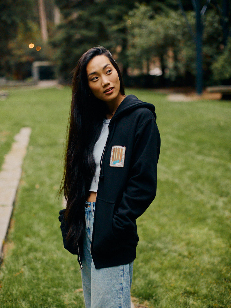 A person with long hair, wearing a Black Muir Woods Patched Zip-Up Hoodie and jeans, stands outdoors on a grass path. They seem free to wander and embrace the natural surroundings.