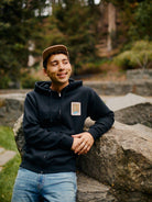 A person in a cap, with a Black Muir Woods Patched Zip-Up Hoodie peeking underneath, leans on a rock, smiling amidst the serene outdoor setting.