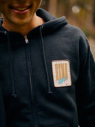 A person in a Black Muir Woods Patched Zip-Up Hoodie, featuring a nature-themed patch with trees and a path, perfectly captures the spirit of "Free To Wander.