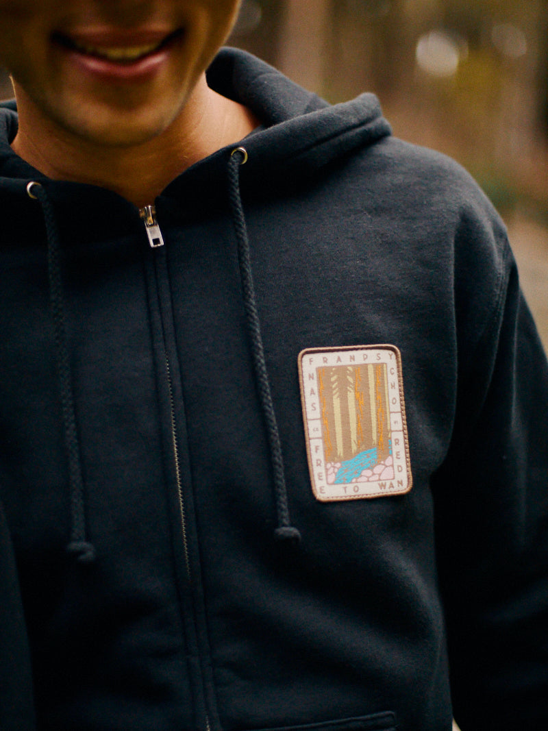 A person in a Black Muir Woods Patched Zip-Up Hoodie, featuring a nature-themed patch with trees and a path, perfectly captures the spirit of "Free To Wander.