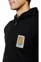 A person wearing the Black Muir Woods Patched Zip-Up Hoodie features a patch of a blue car navigating through a forest, ideal for those seeking freedom in their adventures. Crafted from 100% cotton, it ensures comfort and style for your next journey.