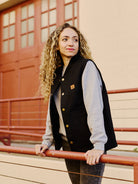 A woman with curly hair in a Leather Patch Workwear Vest, crafted from 100% cotton, leans on a railing, looking to the side.