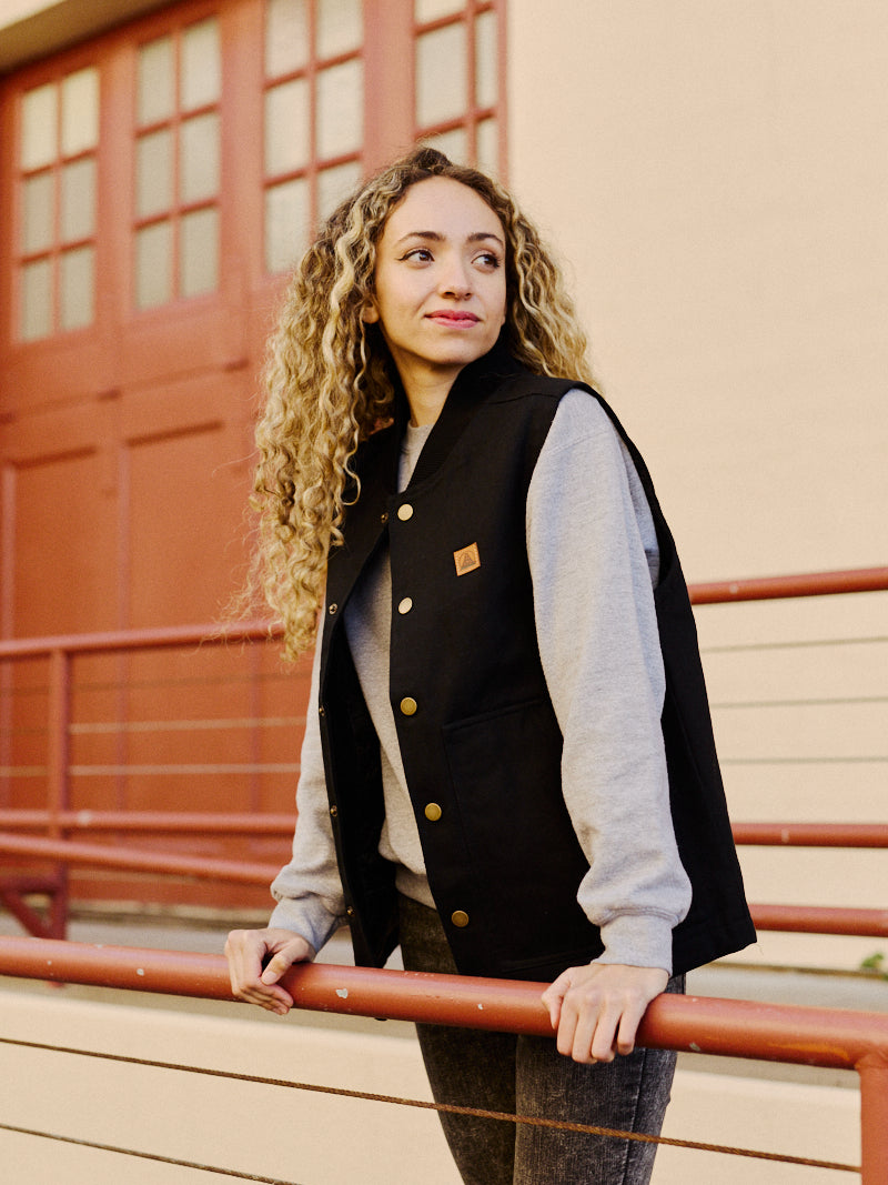 A woman with curly hair in a Leather Patch Workwear Vest, crafted from 100% cotton, leans on a railing, looking to the side.