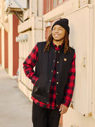 A person in a black beanie and plaid shirt, layered under a Leather Patch Workwear Vest, stands by a building, looking into the distance with a relaxed expression.