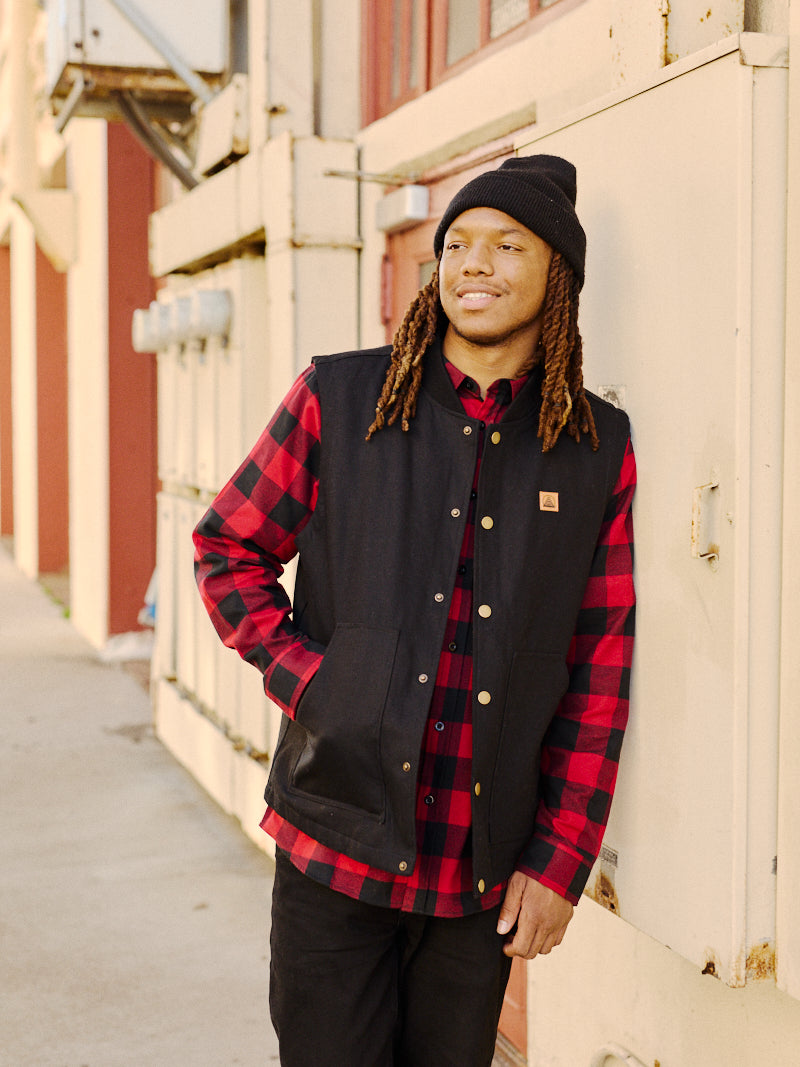 A person in a black beanie and plaid shirt, layered under a Leather Patch Workwear Vest, stands by a building, looking into the distance with a relaxed expression.
