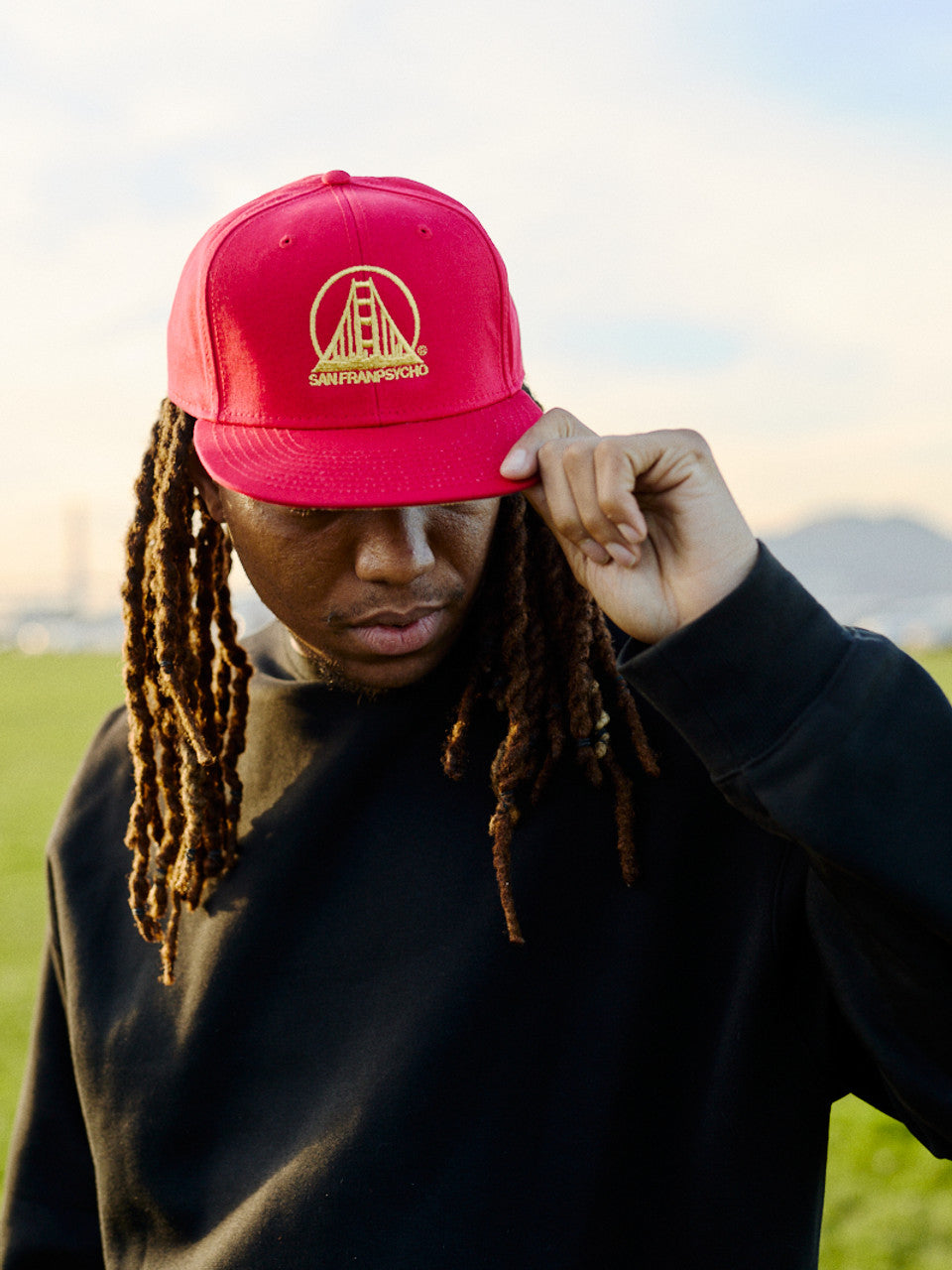 A person with long hair wearing a red Embroidered Logo Snapback - Red & Gold and a black sweatshirt enjoys the outdoors.