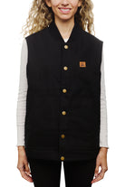 A smiling person wears the sleek Leather Patch Workwear Vest, detailed with gold buttons and front pockets, over a white long-sleeve shirt. Crafted from 100% cotton, this black sleeveless jacket blends style with functionality. Refer to the size guide for the perfect fit.
