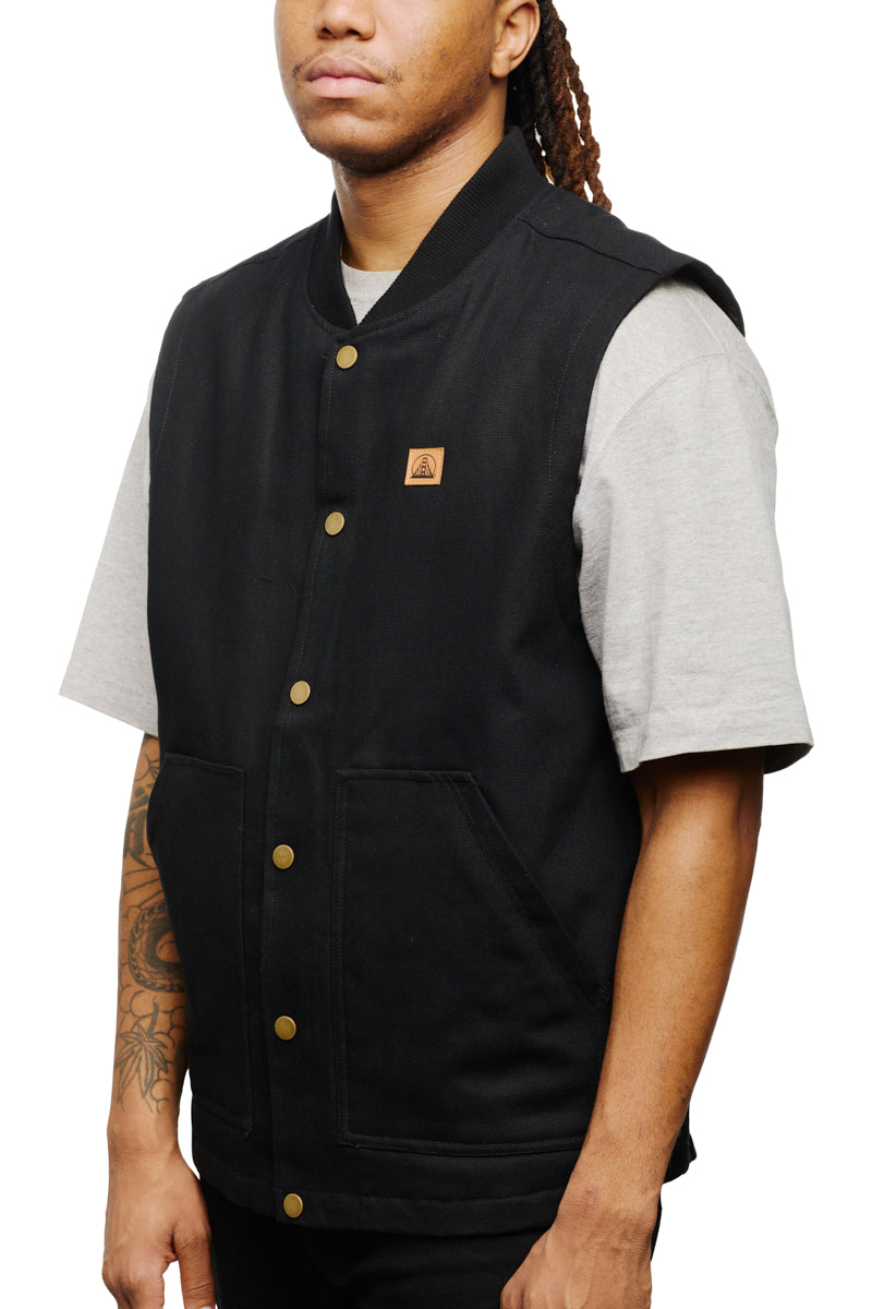 A person stands with hands by their sides, wearing the Leather Patch Workwear Vest—a classic black button-up canvas vest—over a gray T-shirt. Crafted from 100% cotton, this versatile piece combines style and comfort effortlessly. Be sure to check the size guide for the perfect fit.