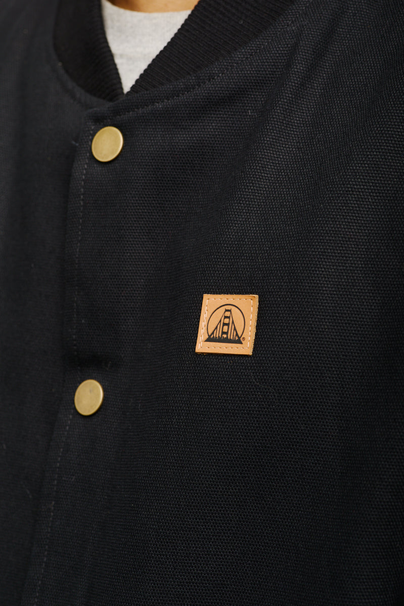 Close-up of the Leather Patch Workwear Vest in black, featuring gold buttons and a small brown square patch adorned with a bridge design, reflecting the rugged style akin to canvas workwear vests.
