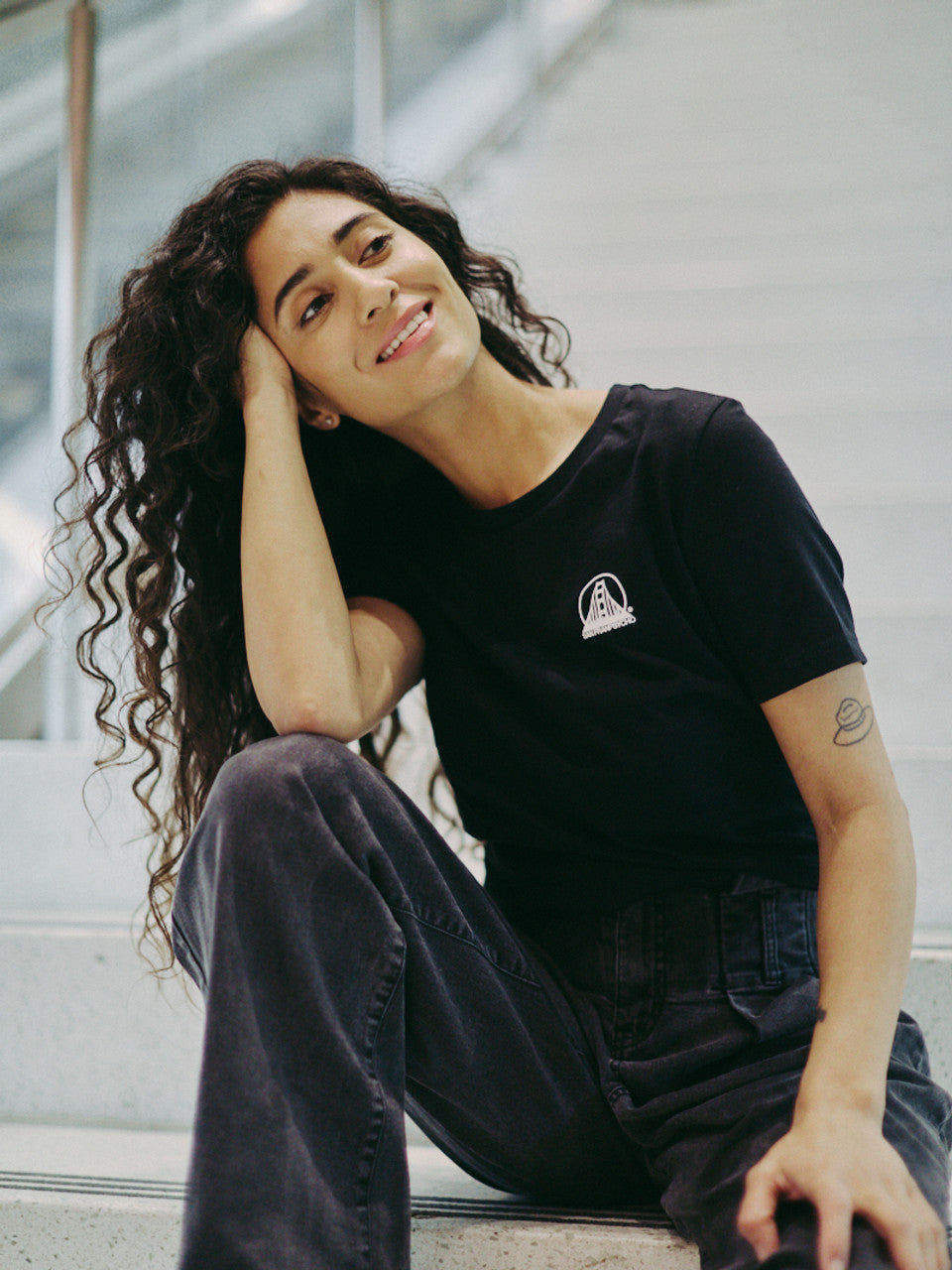 A person with long curly hair, smiling and seated on the stairs, is wearing a Women's Mini Logo Tee in black paired with dark pants.