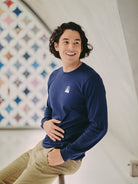 A smiling man with long hair wears a Navy Long Sleeve Mini Logo tee and beige pants while sitting in front of a patterned background.