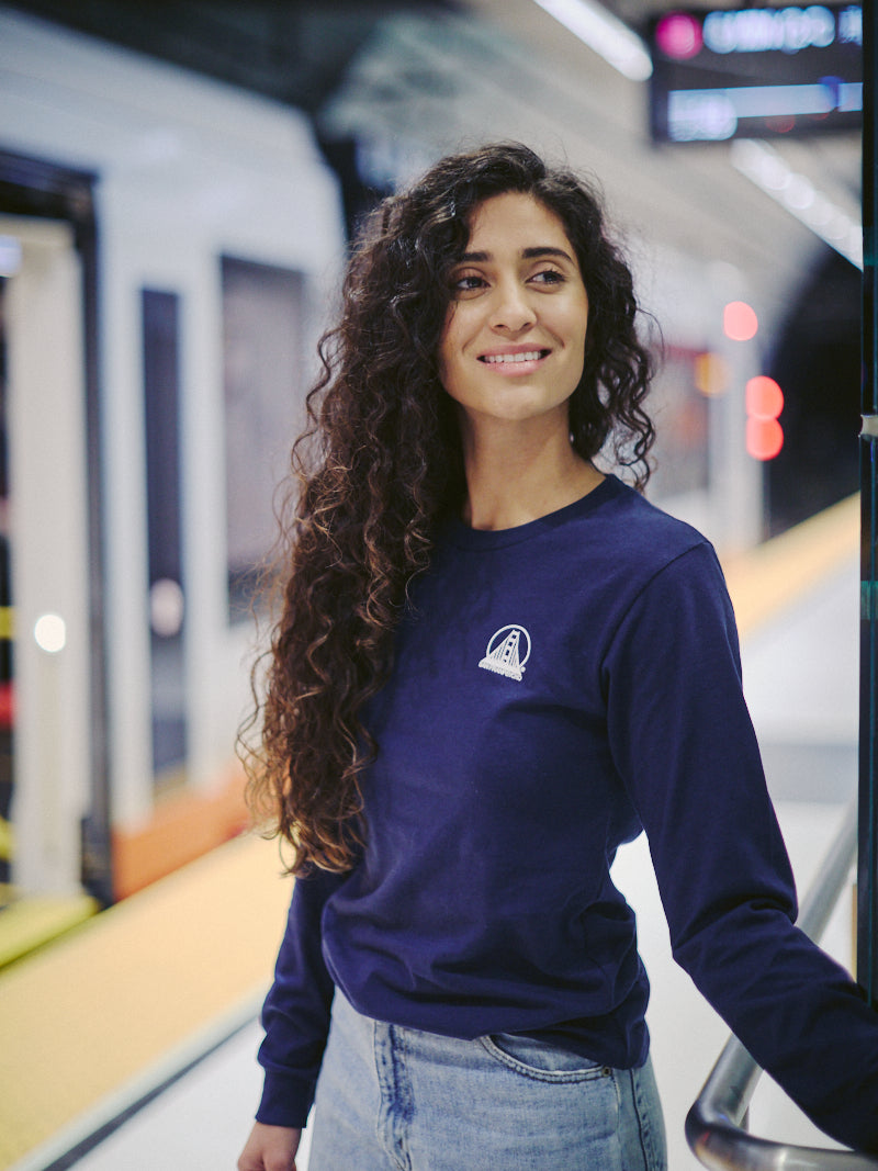 A woman with long curly hair smiles at a train station platform, wearing jeans and a Navy Long Sleeve Mini Logo tee made of soft Airlume cotton.