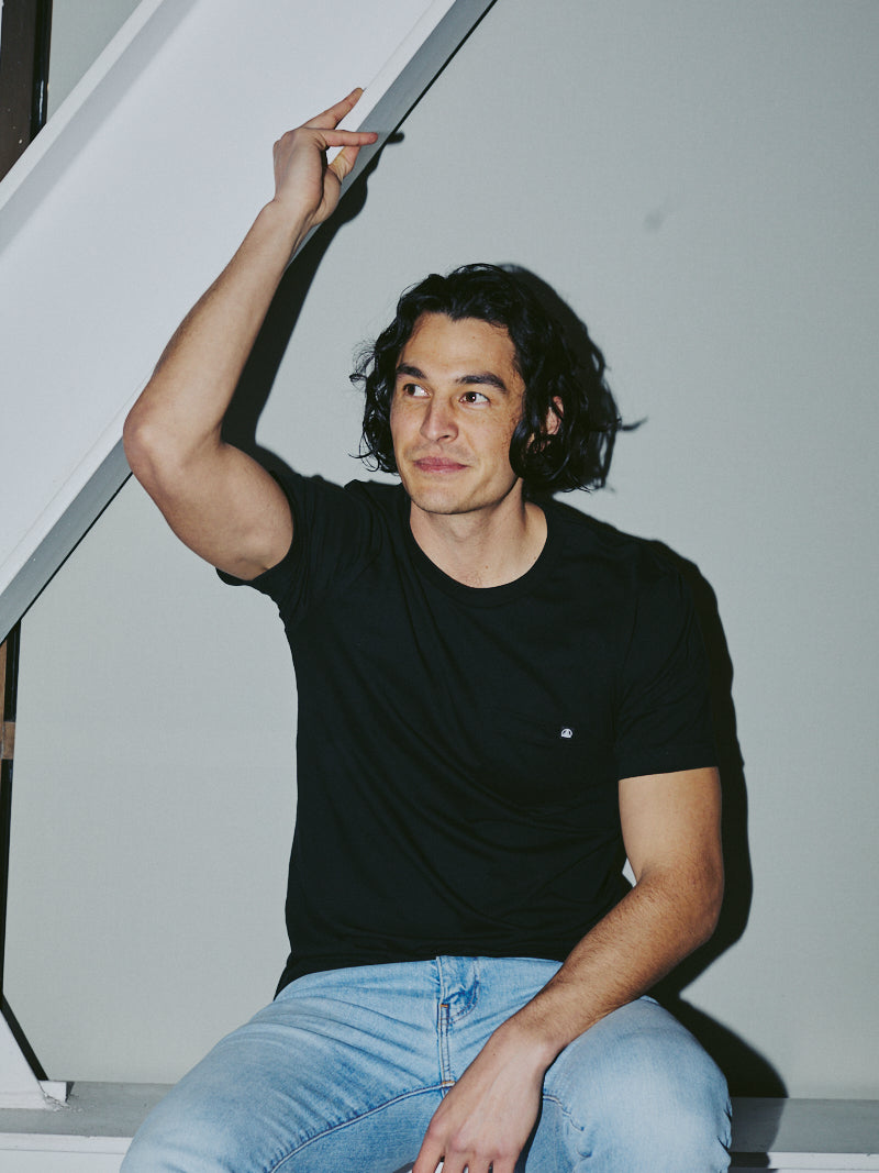 A man in a Black Pocket Tee w/ SFP Clip Label and jeans sits on a staircase, raising his arm with a slight smile.