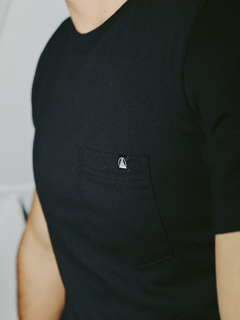 Close-up of a person wearing the Black Pocket Tee w/ SFP Clip Label from the basics line, crafted from soft Airlume cotton, featuring a small logo on the pocket.