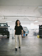 A woman walks confidently in a parking garage, wearing the Black Pocket Tee with SFP Clip Label made from soft Airlume cotton and beige pants, with vehicles in the background.