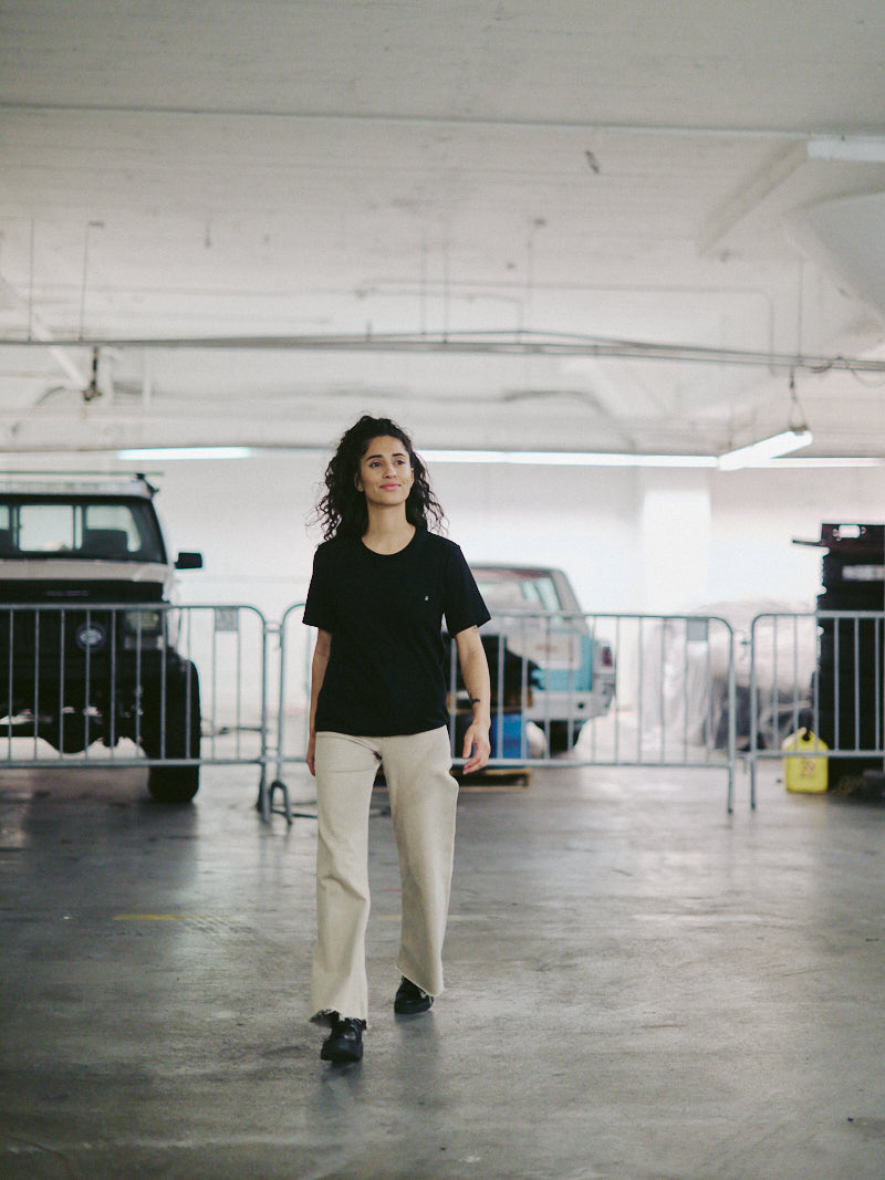 A woman walks confidently in a parking garage, wearing the Black Pocket Tee with SFP Clip Label made from soft Airlume cotton and beige pants, with vehicles in the background.