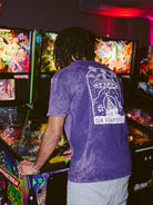 In the dimly lit arcade, a player in a purple Psycho Invader Mineral Wash Tee from the Spring 2023 Collection skillfully navigates the pinball machine, lights reflecting rhythms of concentration.
