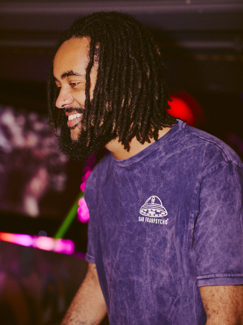 A smiling man with dreadlocks wears a Psycho Invader Mineral Wash Tee, set against a vibrant backdrop with colorful lights, highlighting the Spring 2023 Collection.