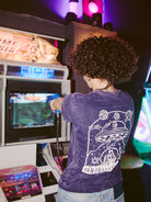 In a dimly lit room, the person immerses themselves in an arcade game, wearing the Psycho Invader Mineral Wash Tee. Their attire subtly echoes the retro-futuristic vibe from the Spring 2023 Collection.