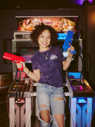 A person is enjoying an arcade shooting game in casual attire, holding two toy guns with a smile. They're wearing the Psycho Invader Mineral Wash Tee from the latest Spring 2023 Collection, adding flair to their gaming experience.
