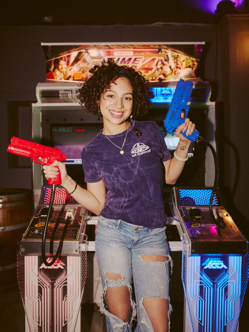 A person is enjoying an arcade shooting game in casual attire, holding two toy guns with a smile. They're wearing the Psycho Invader Mineral Wash Tee from the latest Spring 2023 Collection, adding flair to their gaming experience.