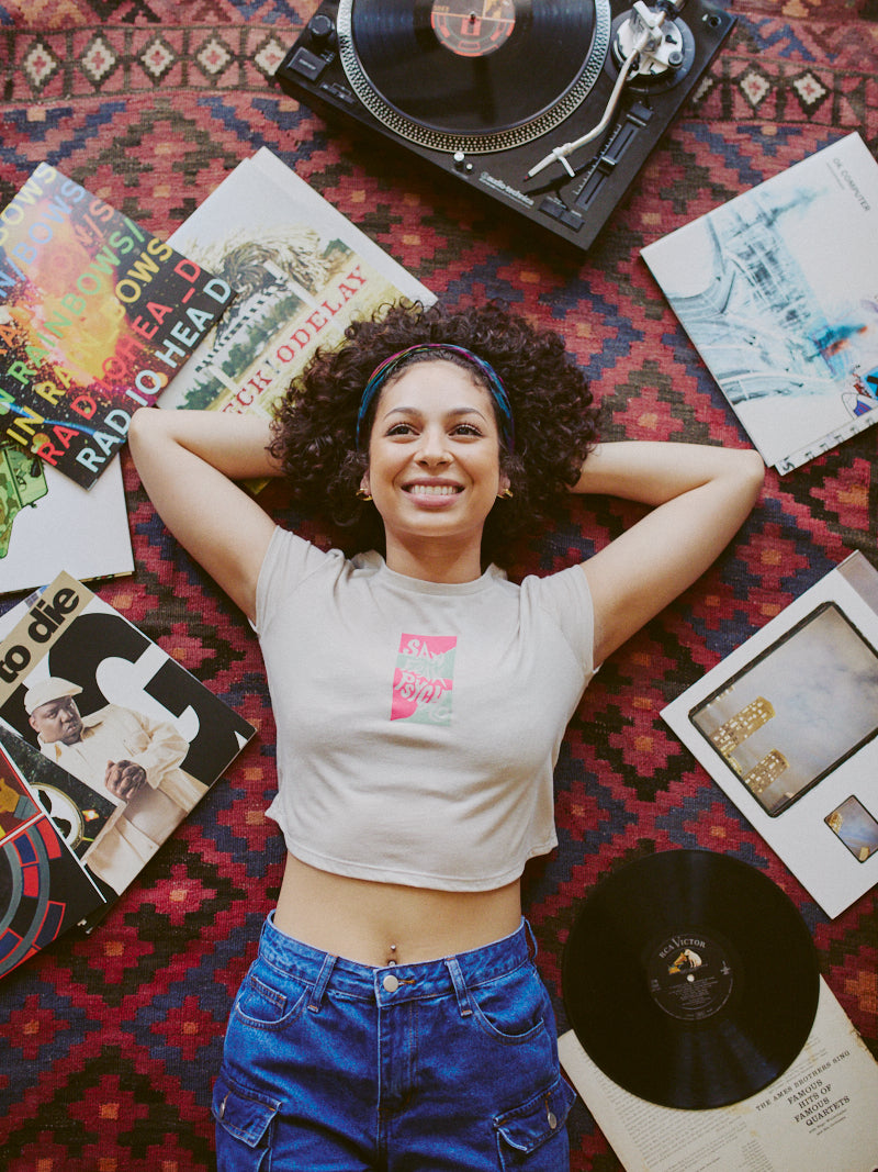 Reclining on a patterned rug, a woman beams at the camera, framed by vinyl records and a turntable. She exudes effortless style in her Trippy Psycho Crop Top from the Spring 2023 Collection.