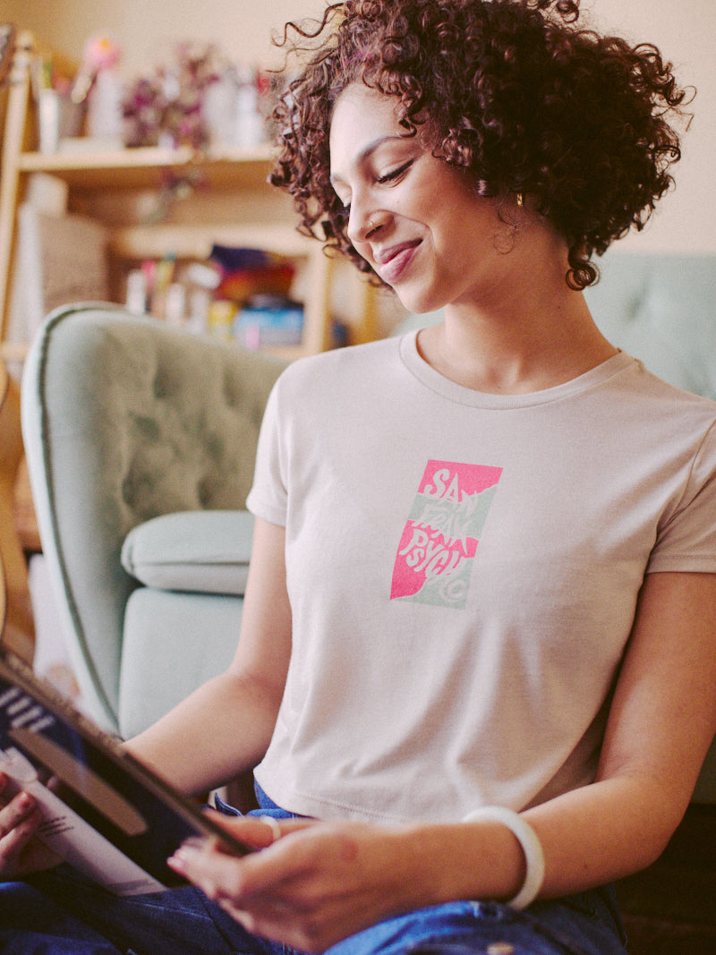 A woman with curly hair, wearing a Trippy Psycho Crop Top, smiles warmly while holding a book in a cozy room. Her hand-printed top from the Spring 2023 Collection adds an artistic touch to the inviting atmosphere.