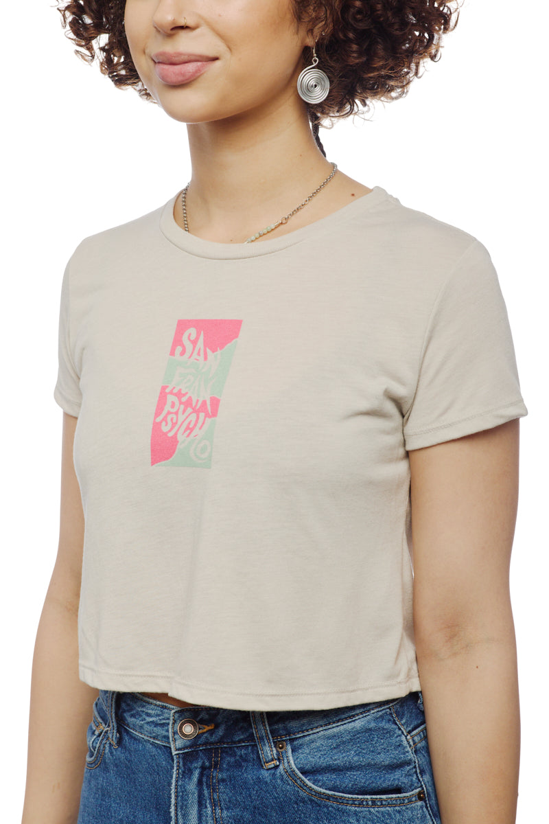 A woman wearing a beige Trippy Psycho Crop Top featuring a graphic design from the Spring 2023 Collection pairs it with blue jeans and has a slight smile.