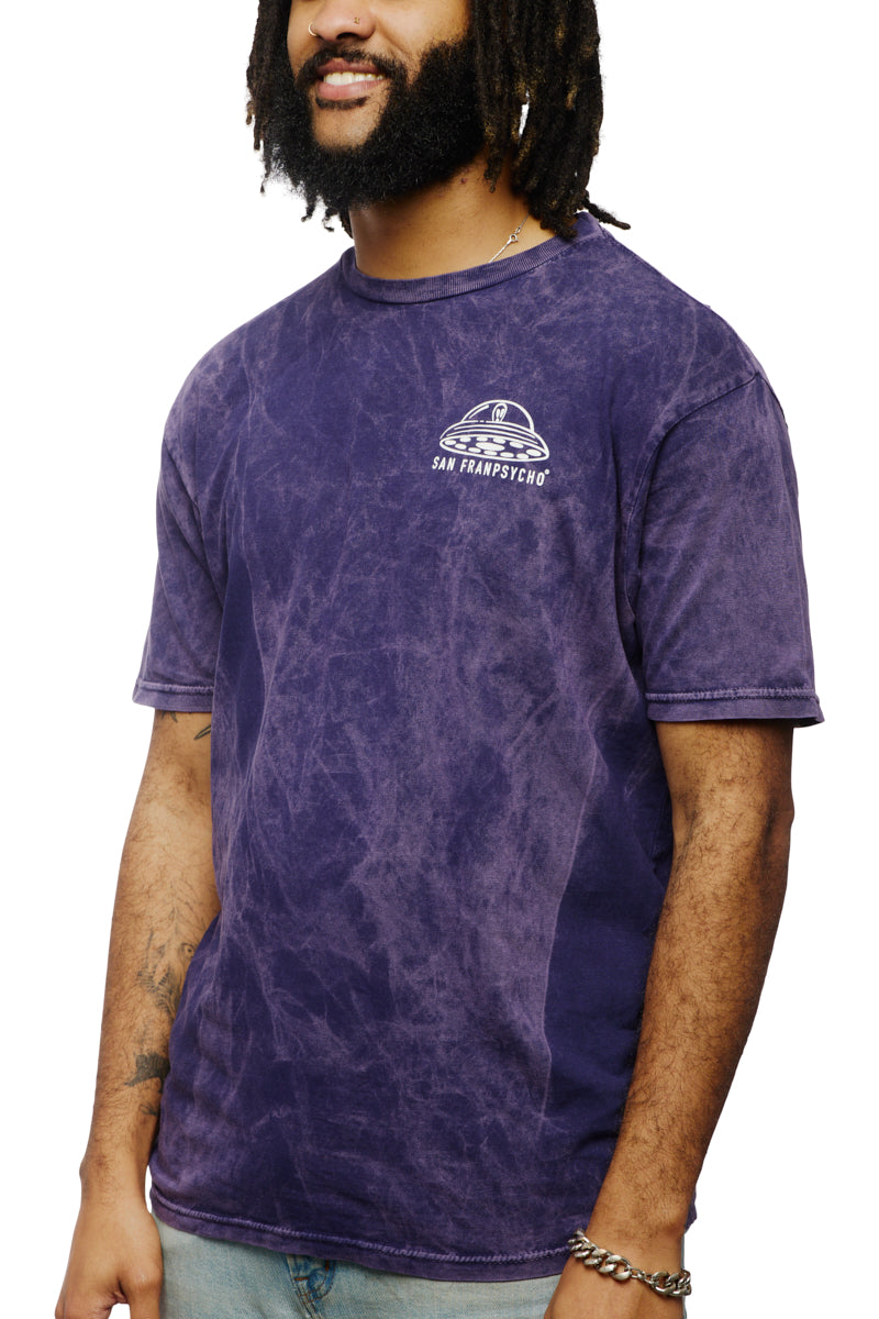 A man wearing the Psycho Invader Mineral Wash Tee from the Spring 2023 Collection, featuring a faded purple color with a bold graphic design of a Psycho Invader, is standing against a white backdrop.