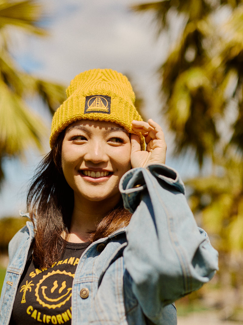 A smiling woman in a mustard lumberjack beanie with the black and yellow SFP logo and a denim jacket enjoys the vibe under palm trees, exuding that unique San Franpsycho charm.
