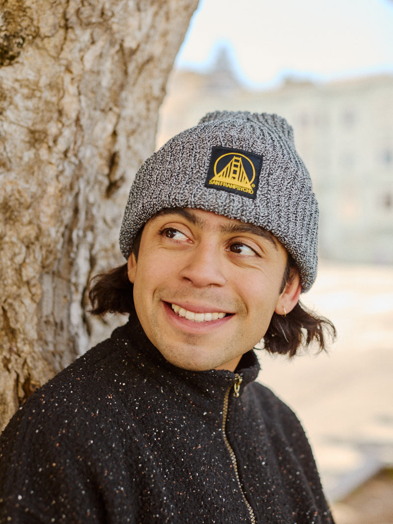 A person wearing a Zebra Lumberjack Beanie with Black/Yellow SFP Logo and a dark jacket smiles while leaning against a tree outdoors.