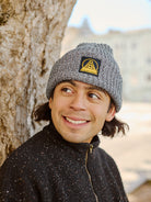 A person wearing a Zebra Lumberjack Beanie with Black/Yellow SFP Logo and a dark jacket smiles while leaning against a tree outdoors.