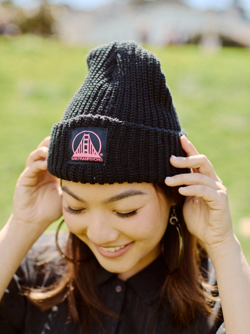 A smiling person adjusts the Black Lumberjack Beanie w/ Black/Pink SFP Logo outdoors on a sunny day.